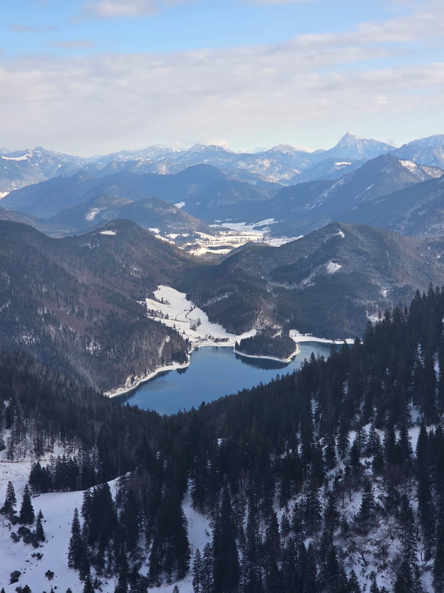 Blick von einem Berg über bewaldete Hänge auf einen See und das Gebirge dahinter